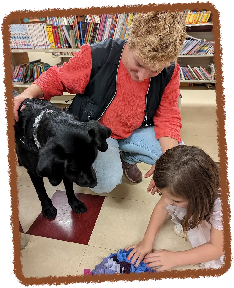 Dans une bibliothèque, Clémentine Reynaud s'occupe d'un chien noir tandis qu'une petite fille est assise à côté d'eux.